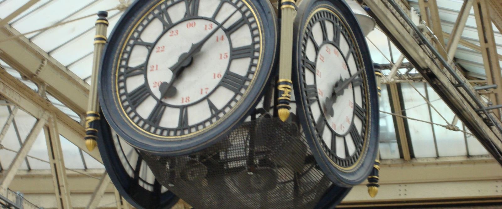 Two faces of a Victorian station clock hanging beneath a glass and iron roof.